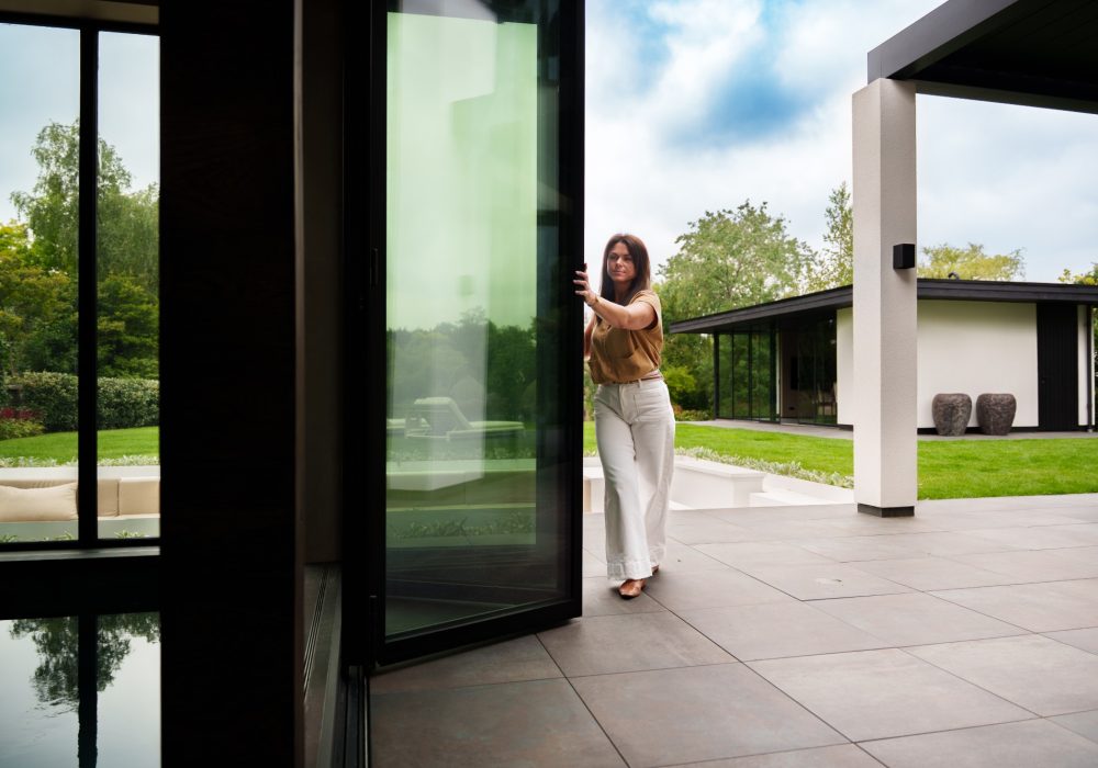 VMR-Vouwwanden-tuin-aanbouw 3 Vrouw die vouwwand aan het openen is waarbij je uitkomt op een deel van het zwembad en de prachtige tuin
